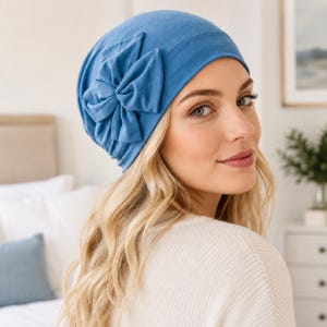 May include: A solid blue beanie hat featuring a large, gathered fabric bow detail on the side. The hat is constructed from a soft, smooth material and fits snugly around the head, providing full coverage with a clean, rounded silhouette.