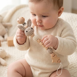 May include: A baby holds a wooden and crochet pacifier clip with a bunny head and the word "BABY" spelled out on wooden beads. The clip has a wooden star and a wooden pacifier holder. The baby is wearing a cream-colored onesie.