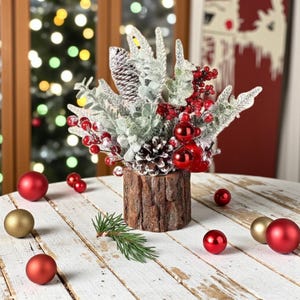 Puede incluir: Un centro de mesa navideño festivo en un recipiente de madera rústica. Presenta vegetación helada, bayas rojas, piñas y adornos rojos. Varios adornos rojos y dorados están esparcidos sobre la superficie de madera blanca.