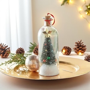 Puede incluir: Una botella de vidrio transparente con tapón de corcho y cinta roja contiene un árbol de Navidad en miniatura con adornos coloridos y una estrella dorada. La botella está sobre un plato dorado, rodeada de piñas y vegetación.