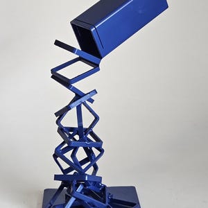 May include: A blue metal sculpture featuring a series of interconnected square frames stacked in a precarious, leaning formation, topped with a solid rectangular block, all mounted on a flat, square blue metal base.