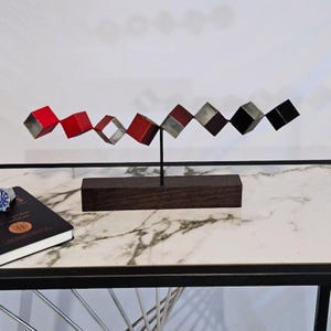 May include: Abstract metal sculpture featuring a series of connected, open cubes in red, silver, brown, and black. The sculpture is mounted on a dark wooden base and supported by a thin, black metal rod. The artwork is displayed on a marble-patterned surface.