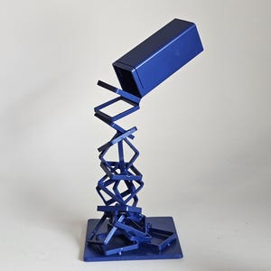 May include: A dark blue metal sculpture featuring a solid rectangular block suspended above a square base. The block is connected to the base by a series of interconnected, tilted square metal frames that create a cascading, gravity-defying effect.