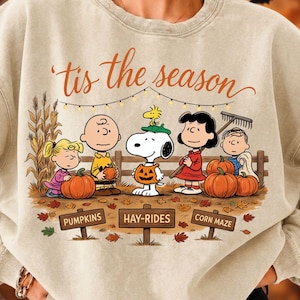 May include: A beige crewneck sweatshirt featuring a graphic of the Peanuts characters in a pumpkin patch. The text 'tis the season' appears above, with signs below reading 'PUMPKINS', 'HAY-RIDES', and 'CORN MAZE' amidst fallen autumn leaves.