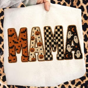 May include: A white sweatshirt featuring the word MAMA in large, textured, embroidered letters. Each letter has a unique pattern: orange with black bats, cream with orange pumpkins, a black and white checkerboard, and black with white ghosts.