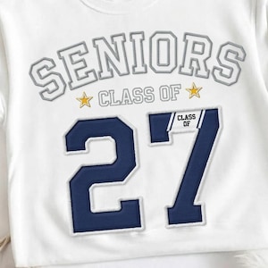 Op de afbeelding: Een wit sweatshirt met ronde hals voorzien van het woord SENIORS in grijze blokletters, gevolgd door de tekst CLASS OF met twee gele sterren. Daaronder staan grote marineblauwe cijfers 27, met de tekst CLASS OF op het cijfer 7.