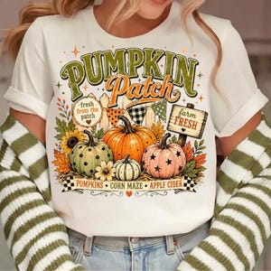 Op de afbeelding: Een wit t-shirt met een grafische print met de tekst PUMPKIN Patch, fresh from the patch en farm fresh. Het ontwerp bevat drie pompoenen in groen, oranje en roze, zonnebloemen, herfstbladeren en een houten hek met vlaggetjes.