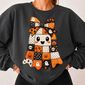 Pode incluir: Uma camisola cinzenta escura de gola redonda com um grande gráfico central de um fantasma com padrão de patchwork. O fantasma está adornado com um laço grande e decorado com abóboras cor de laranja, caveiras brancas, fantasmas pequenos e pontos pretos.