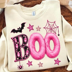 May include: A cream-colored crewneck sweatshirt featuring the word BOO in large letters. The B is black with pink gemstone accents, while the two Os are textured pink faux fur. The design includes a black glitter bat, a pink spiderweb, a small spider, and pink stars.