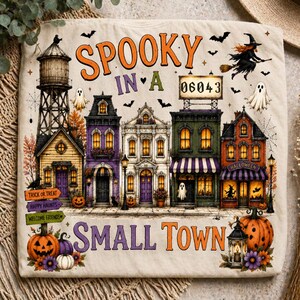 Könnte beinhalten: Ein cremefarbener Stoff mit einer Halloween-Illustration einer Kleinstadt mit Wasserturm, Geschäften und Häusern. Der Text lautet "SPOOKY IN A SMALL TOWN" mit "06043" auf einem Schild. Enthält Geister, eine Hexe, Kürbisse und Wegweiser.