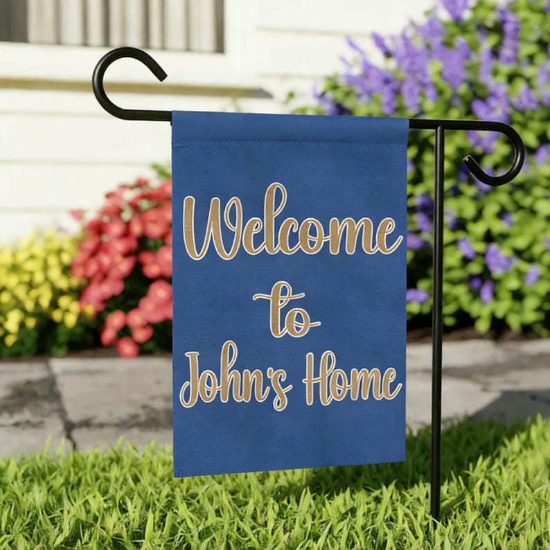 May include: A blue garden flag with the words "Welcome to John's Home" in gold script. The flag is displayed on a black metal stand in a grassy yard, with colorful flowers in the background.