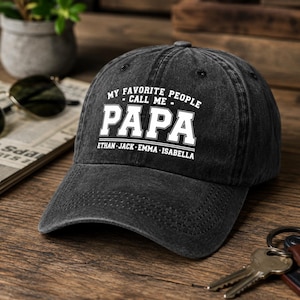 May include: A black, distressed baseball cap with the text "MY FAVORITE PEOPLE CALL ME PAPA" in white block letters. Below the word "PAPA" are the names "ETHAN JACK EMMA ISABELLA". The cap is sitting on a wooden surface.