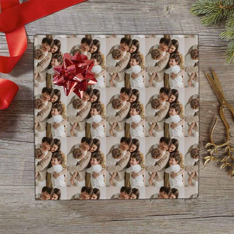 May include: Wrapping paper featuring a repeating pattern of a family embracing. A large red bow sits on top of the paper. The scene includes a red ribbon, gold scissors, and a sprig of evergreen on a wooden surface.