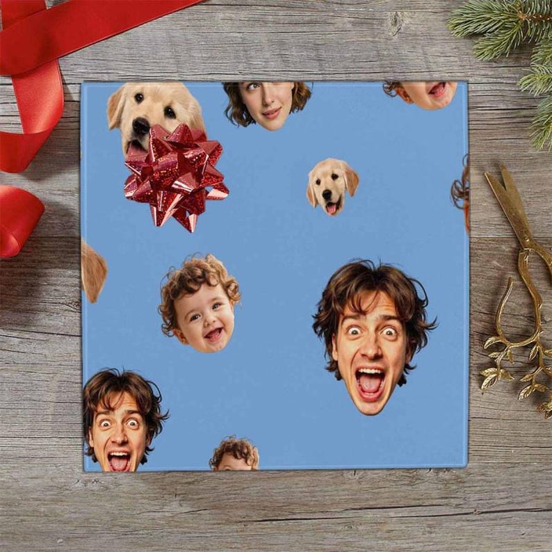 May include: Blue wrapping paper with a repeating pattern of smiling faces of people and dogs. A red bow is on the top left. The paper is on a wooden surface with a red ribbon and scissors.