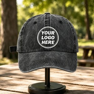 May include: A charcoal grey acid-wash baseball cap featuring a white circular graphic on the front panel with the text YOUR LOGO HERE centered inside. The cap has a curved brim, stitched eyelets, and a fabric strap closure on the side.