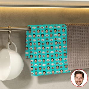 May include: A teal kitchen towel featuring a repeating pattern of a man's face and small white dogs. The towel is hanging on a metal bar next to a brown waffle-weave towel and a white mug.