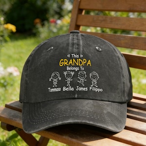 May include: A black baseball cap with the text "This Grandpa Belongs To" and the names Tommaso Bella James Filippo in white. The word "Grandpa" is in yellow. The cap is sitting on a wooden bench.