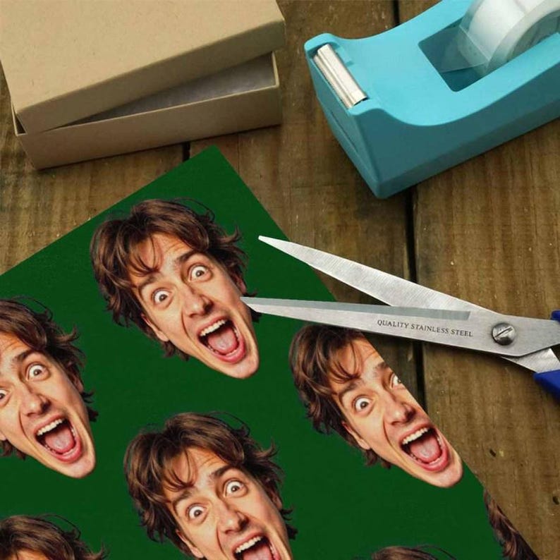 May include: A green sheet with multiple images of a man's face with an open mouth and wide eyes. Scissors are cutting the sheet. A blue tape dispenser and a brown box are in the background.