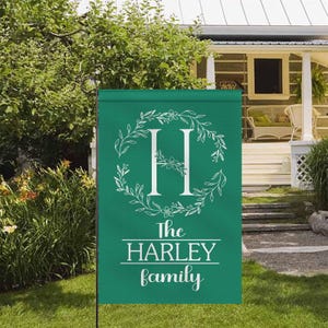 May include: A green garden flag with a white floral wreath design around the letter "H". The text "The HARLEY family" is displayed in white below the letter. The flag is displayed outdoors in front of a house.
