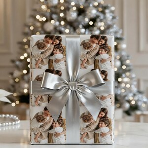 May include: A wrapped gift box featuring a family photo repeated across the wrapping paper. The gift is tied with a wide silver satin ribbon and bow. The background includes a blurred, decorated Christmas tree.