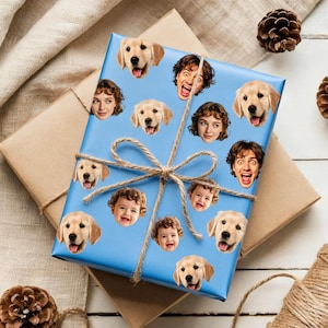 May include: A wrapped gift with light blue paper featuring a pattern of golden retriever faces, smiling faces, and baby faces. The gift is tied with twine and sits on top of a brown paper-wrapped gift.