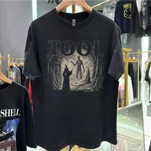 May include: Black t-shirt featuring the band name "TOOL" in large, gray block letters above a detailed graphic. The graphic depicts a figure in a robe and a skeletal figure in a cave setting. The shirt is hanging on a wooden hanger.