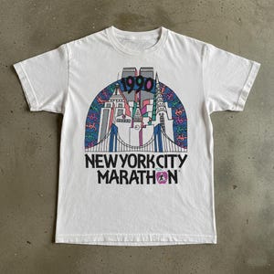 May include: White t-shirt with a graphic depicting New York City landmarks, bridges, and the text "1990" and "NEW YORK CITY MARATHON". The design incorporates blue, pink, green, and purple, with a runner silhouette.
