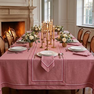 May include: A formal dining table set for a special occasion. The table is covered with a dusty rose tablecloth and runner, set with white plates, silverware, and napkins. Gold candlesticks with lit candles and floral arrangements complete the elegant setting.