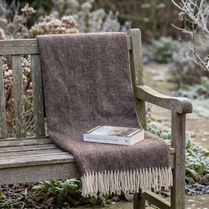 May include: A brown herringbone-patterned throw blanket with cream-colored fringe is draped over a wooden bench. A book titled "A Tale of Two Cities" rests on the blanket, suggesting a cozy reading nook. The scene is set outdoors.
