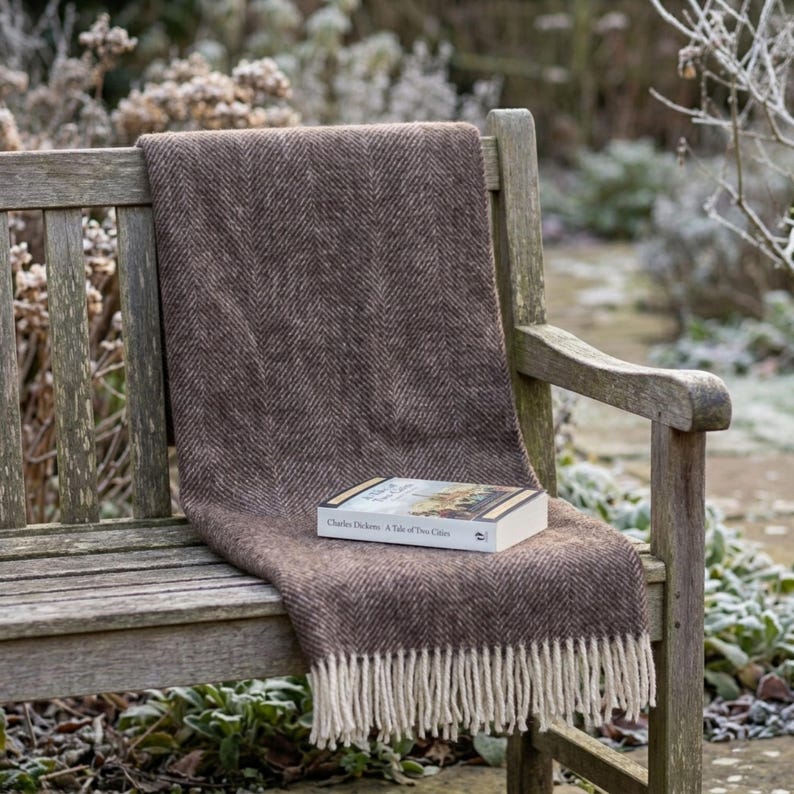 May include: A brown herringbone-patterned throw blanket with cream-colored fringe is draped over a wooden bench. A book titled "A Tale of Two Cities" rests on the blanket, suggesting a cozy reading nook. The scene is set outdoors.