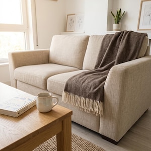 May include: A beige two-seater sofa with a brown fringed throw blanket. A wooden coffee table holds a mug and a book titled "HYGGE". The room has framed botanical prints and a small rug.
