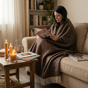 May include: A woman wrapped in a brown herringbone blanket reads a TIME magazine on a beige sofa. A wooden side table holds lit candles, a mug, and magazines. A bookshelf is visible in the background.