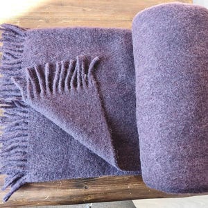 May include: A deep purple throw blanket with a textured, felted appearance. The blanket is partially rolled into a cylinder shape, with the remaining portion laid flat, showcasing a series of fringe tassels along the edge of the fabric.