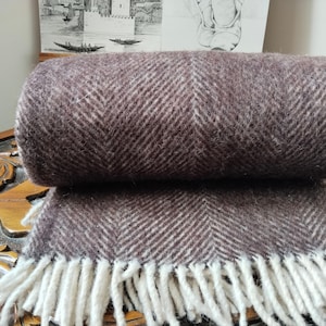 May include: A rolled-up brown and white herringbone pattern throw blanket with a fringe. The blanket is made of a soft, textured material. The fringe is white. The blanket is resting on a wooden surface.