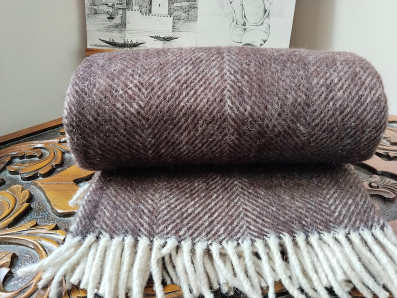 May include: A rolled-up brown and white herringbone pattern throw blanket with a fringe. The blanket is made of a soft, textured material. The fringe is white. The blanket is resting on a wooden surface.