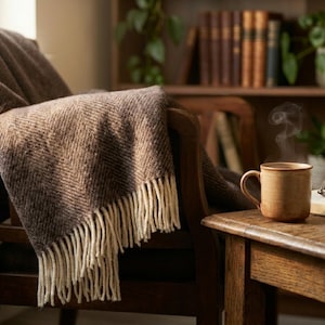 May include: A cozy scene featuring a brown blanket draped over a wooden chair. A mug of steaming coffee sits on a side table next to an open book and glasses. The room has a warm, inviting atmosphere.