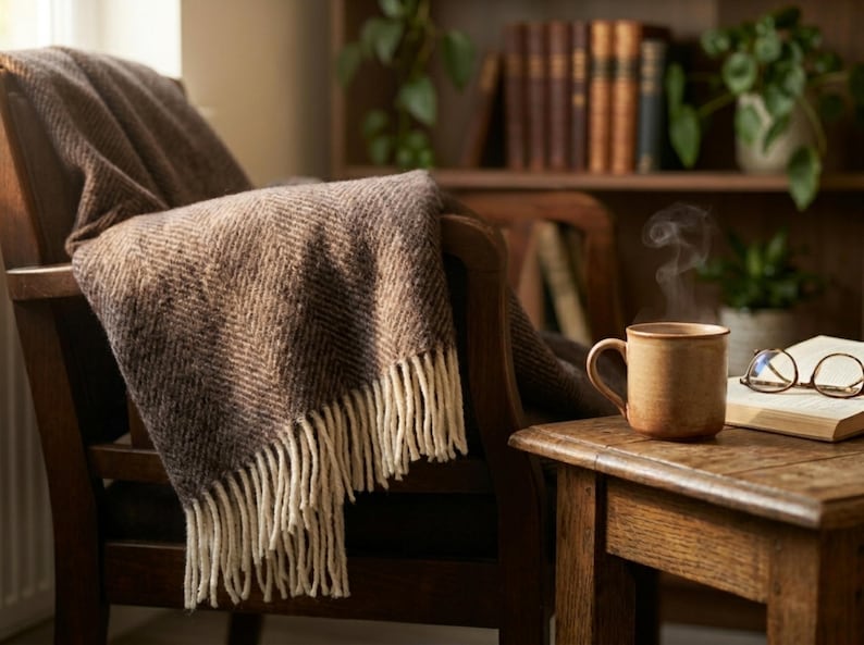 May include: A cozy scene featuring a brown blanket draped over a wooden chair. A mug of steaming coffee sits on a side table next to an open book and glasses. The room has a warm, inviting atmosphere.
