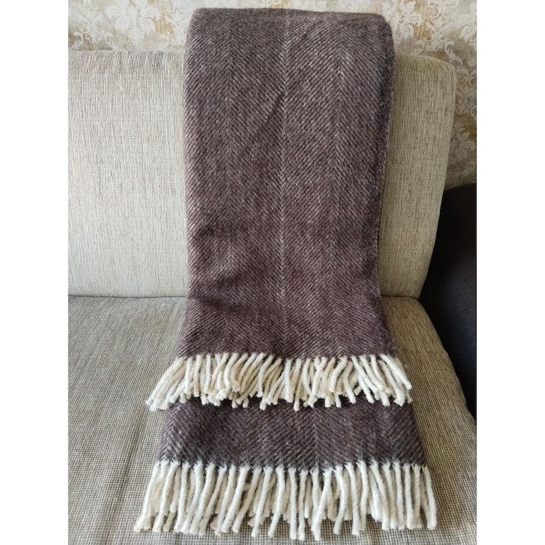 May include: A brown herringbone-patterned throw blanket with cream-colored fringe. The blanket is folded over a light-colored sofa. The throw has a textured appearance and is made of a soft material.