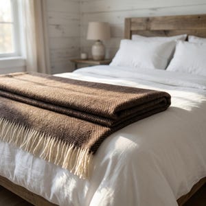 May include: A folded brown and cream-colored blanket with a herringbone pattern rests on a white bed. The blanket has a fringe along the bottom edge. The bedroom has a rustic wooden headboard and a white wall.