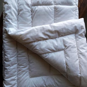 May include: A white quilted comforter featuring a grid-like stitched pattern across the surface. The bedding is folded over at the top corner, revealing the soft, layered construction and the smooth, solid white fabric texture of the material.