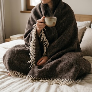 May include: A person wrapped in a brown herringbone patterned blanket with cream-colored fringe, holding a ceramic mug. The blanket covers the person sitting on a bed with beige linens and pillows. A cozy, warm scene.