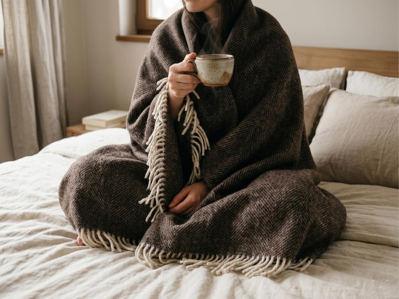 May include: A person wrapped in a brown herringbone patterned blanket with cream-colored fringe, holding a ceramic mug. The blanket covers the person sitting on a bed with beige linens and pillows. A cozy, warm scene.