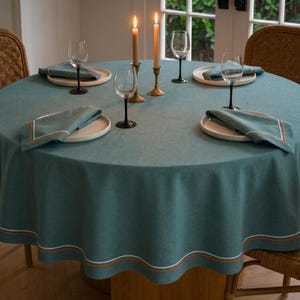 Puede incluir: Un mantel redondo color turquesa con borde festoneado, preparado para una cena. La mesa está puesta con platos, servilletas y copas de vino. Dos velas encendidas en candelabros de latón añaden un brillo cálido. Sillas de mimbre rodean la mesa.
