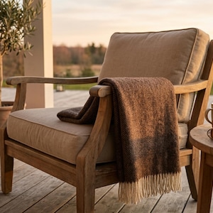May include: A wooden outdoor armchair with a beige cushion and a brown blanket draped over the arm. A small side table holds a mug and a book. An olive tree in a terracotta pot is in the background.
