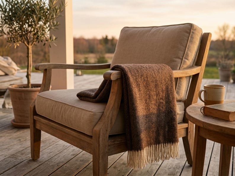 May include: A wooden outdoor armchair with a beige cushion and a brown blanket draped over the arm. A small side table holds a mug and a book. An olive tree in a terracotta pot is in the background.