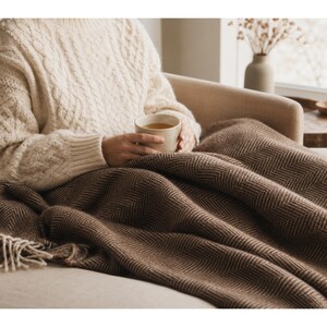 May include: A cozy scene featuring a cream-colored cable-knit sweater and a brown herringbone blanket. The blanket is draped over a beige sofa. A person is holding a mug. The scene evokes warmth and comfort.