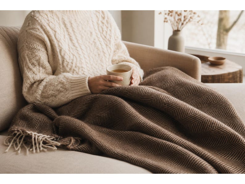 May include: A cozy scene featuring a cream-colored cable-knit sweater and a brown herringbone blanket. The blanket is draped over a beige sofa. A person is holding a mug. The scene evokes warmth and comfort.