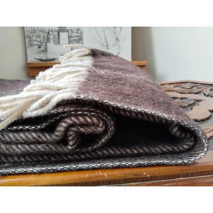 May include: A folded brown and cream-colored blanket with a fringed edge. The blanket has a herringbone pattern and is made of a thick, woven material. The blanket is folded over, showing the different layers and textures.