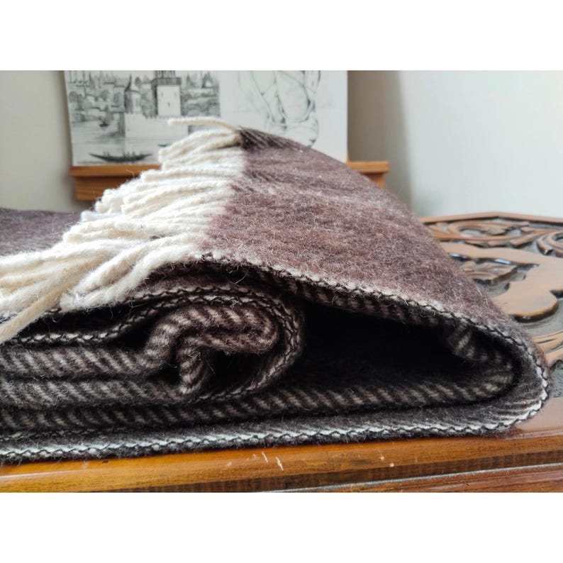May include: A folded brown and cream-colored blanket with a fringed edge. The blanket has a herringbone pattern and is made of a thick, woven material. The blanket is folded over, showing the different layers and textures.