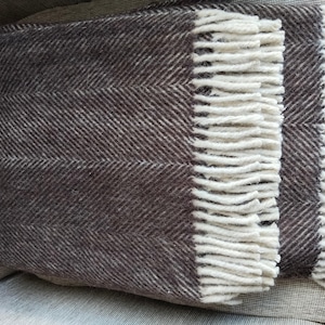 May include: A folded brown and white herringbone pattern throw blanket with cream-colored fringe. The blanket is made of a soft, textured material and is folded neatly. The fringe adds a decorative touch to the blanket's edge.